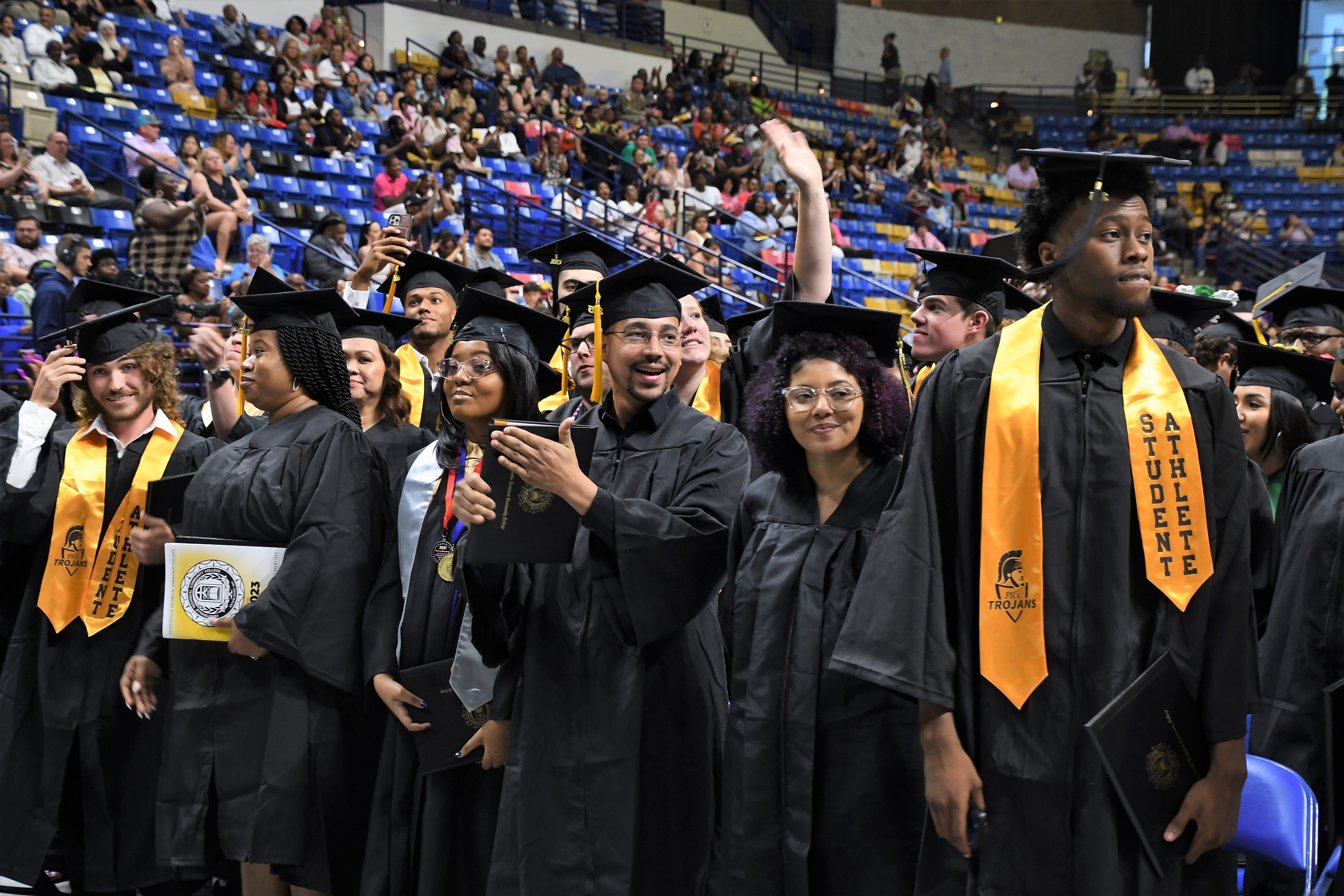 Graduates standing in front of rows of chairs stand and clap.