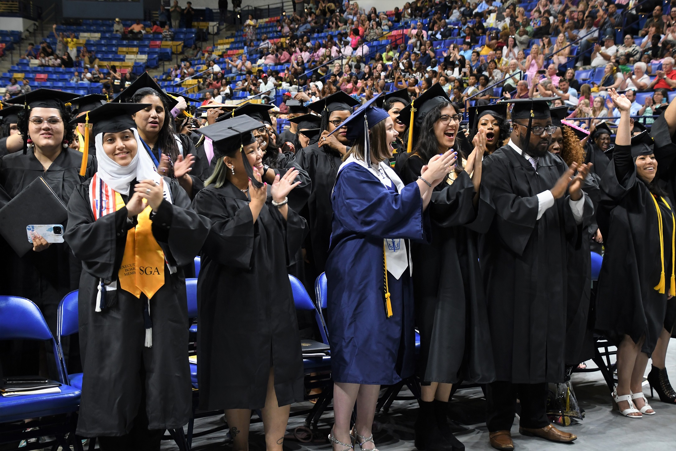 Graduates stand in front of the chair and lift their hands to reach for their tassels.