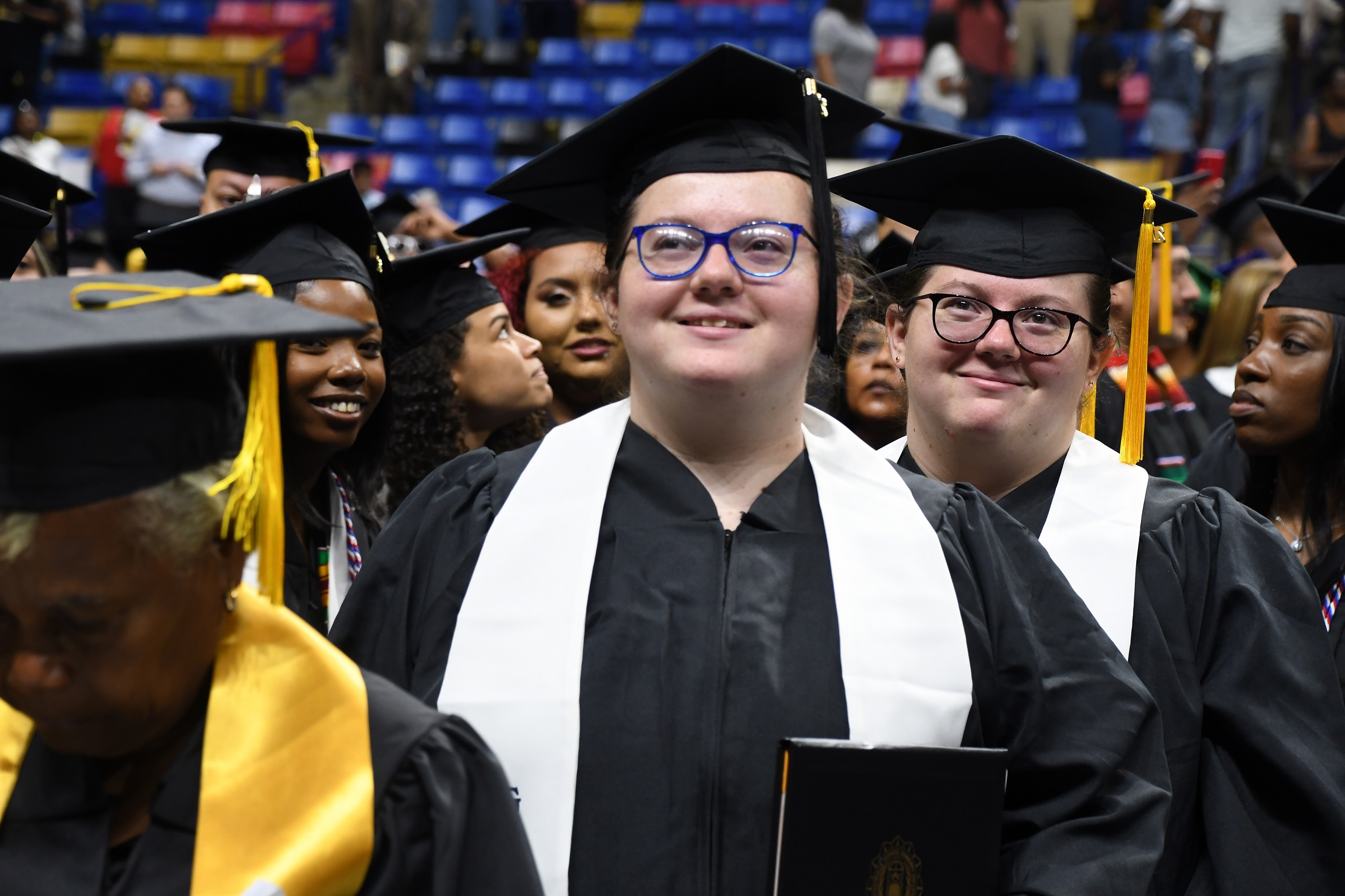 Two graduates, both wearing glasses and white stoles, smile at the camera as they walk.