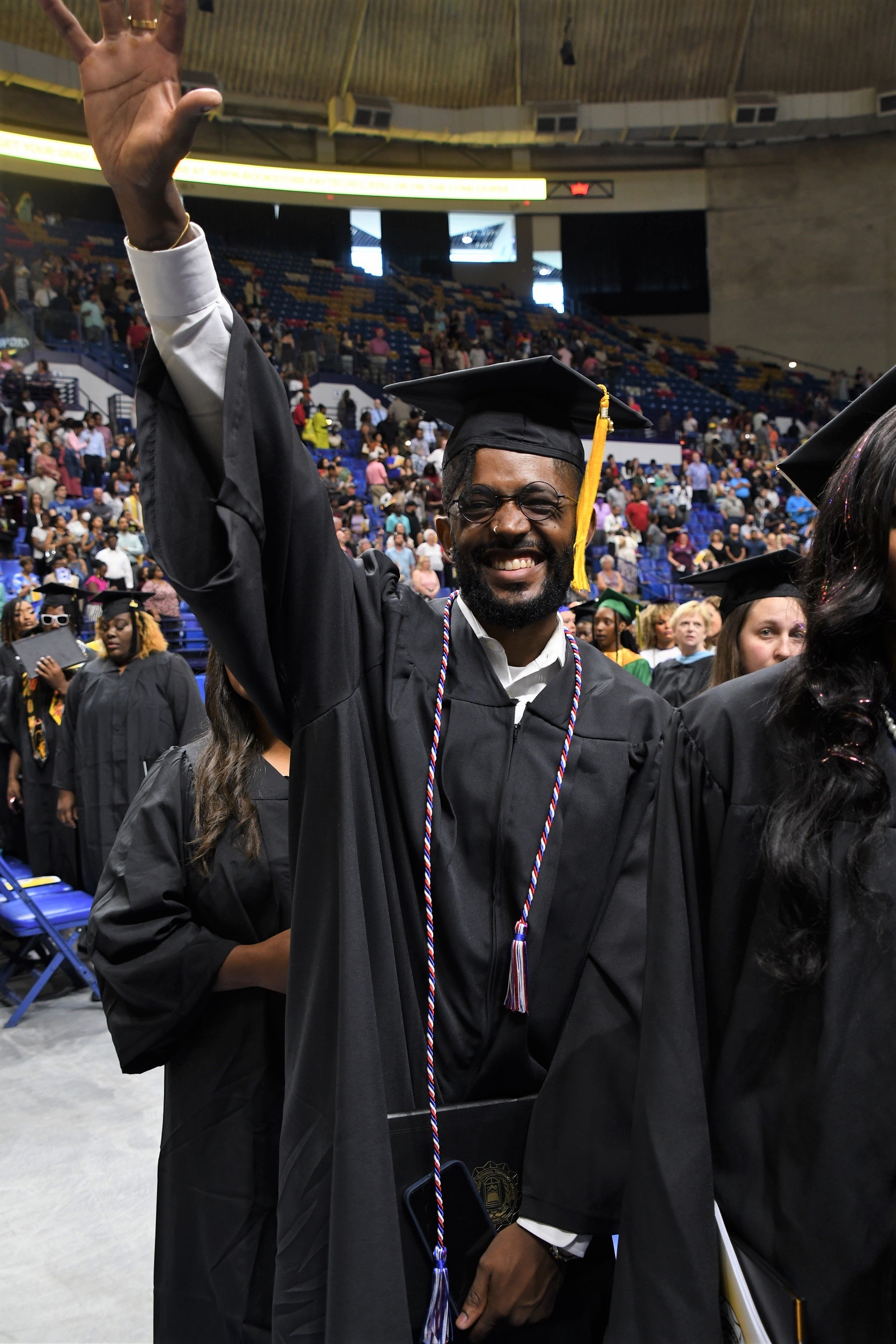 A graduate wearing a red, white and blue cord, smiles and raises his hand in a wave at the camera.