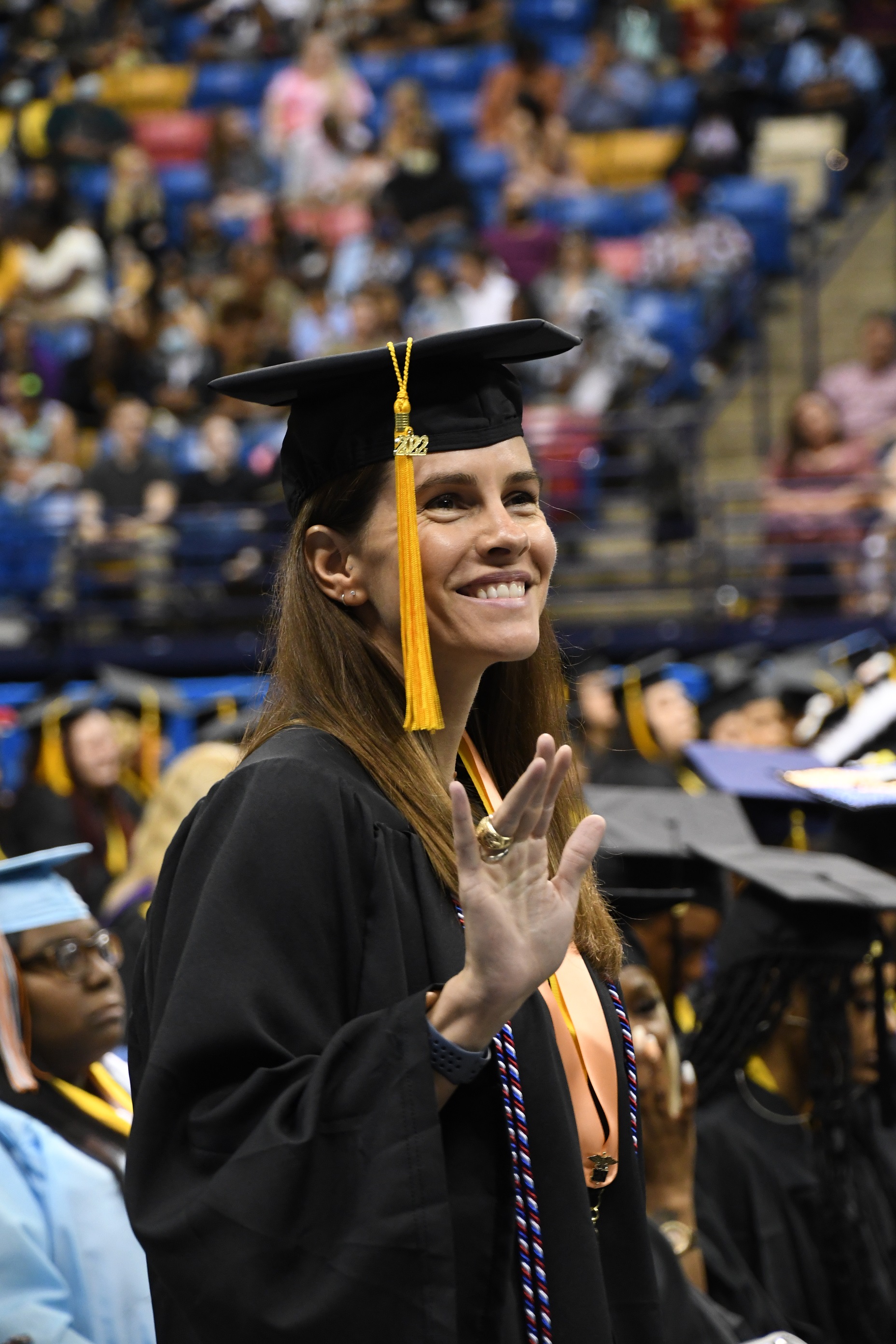 A graduates smiles and waves with a slightly raised hand to the crowd.