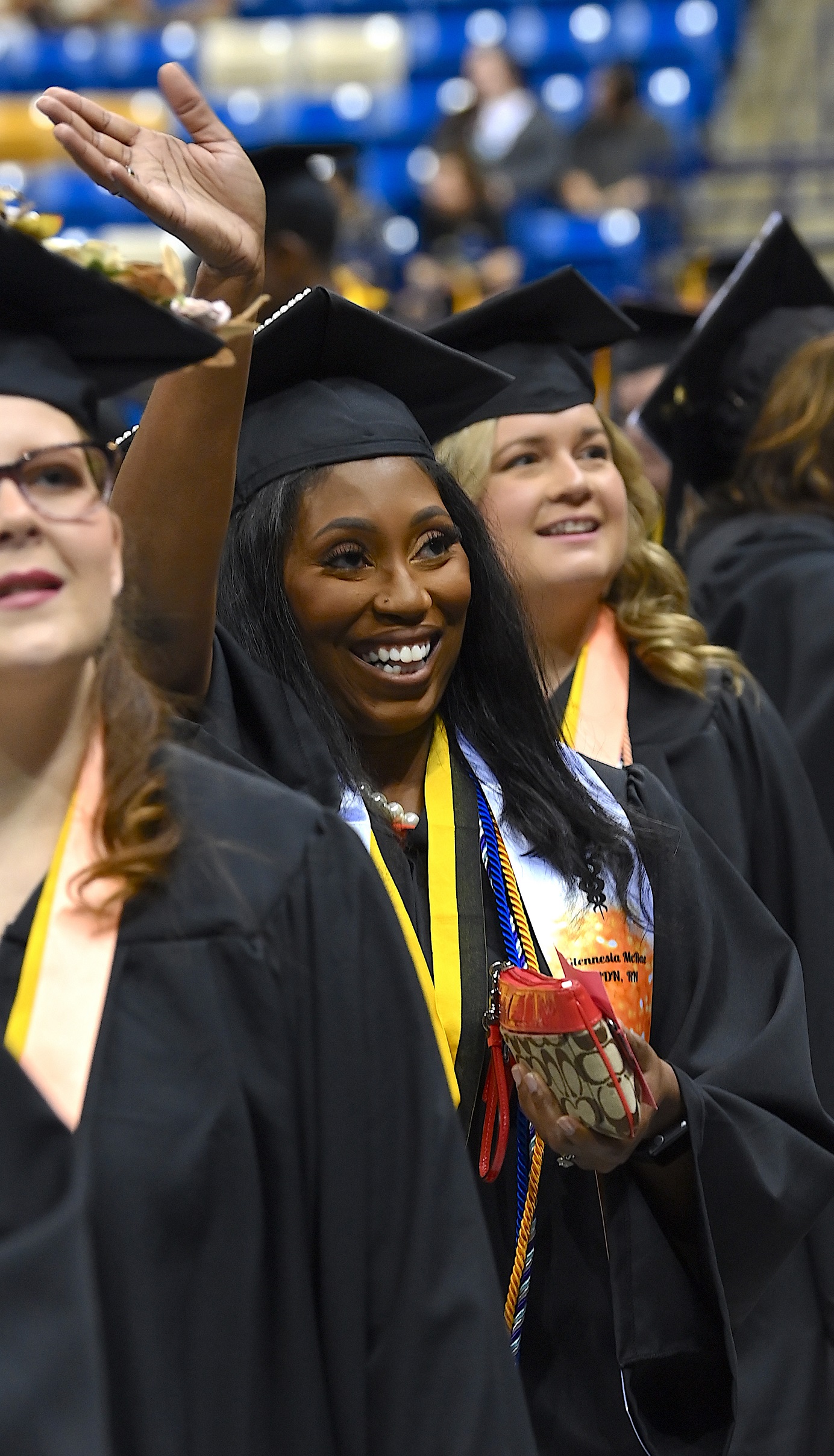 A graduate wearing a custom blue and gold stole smiles and holds her hand high to wave to the crowd.