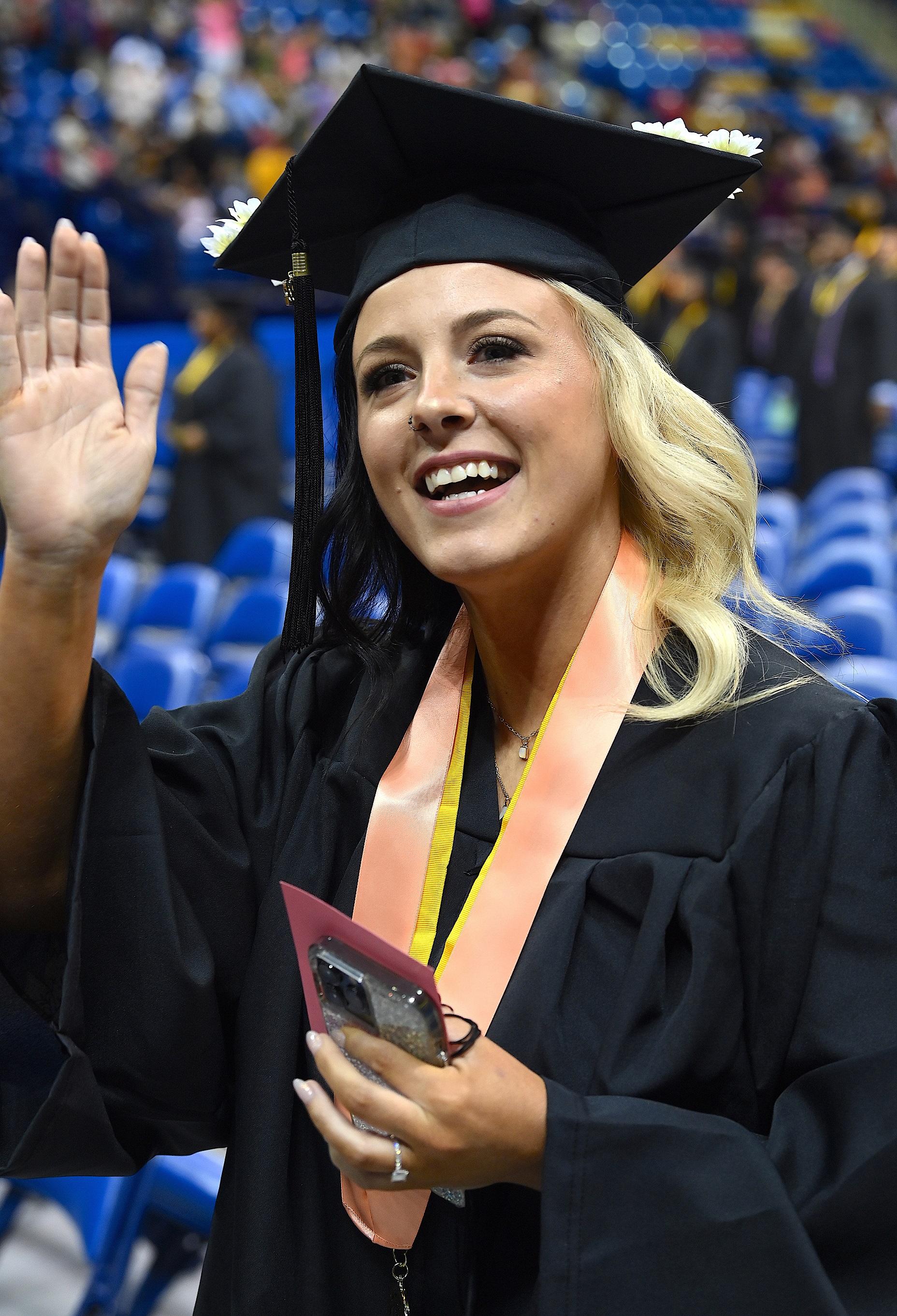 A graduate wearing a peach ribbon around her neck waves to the crowd.