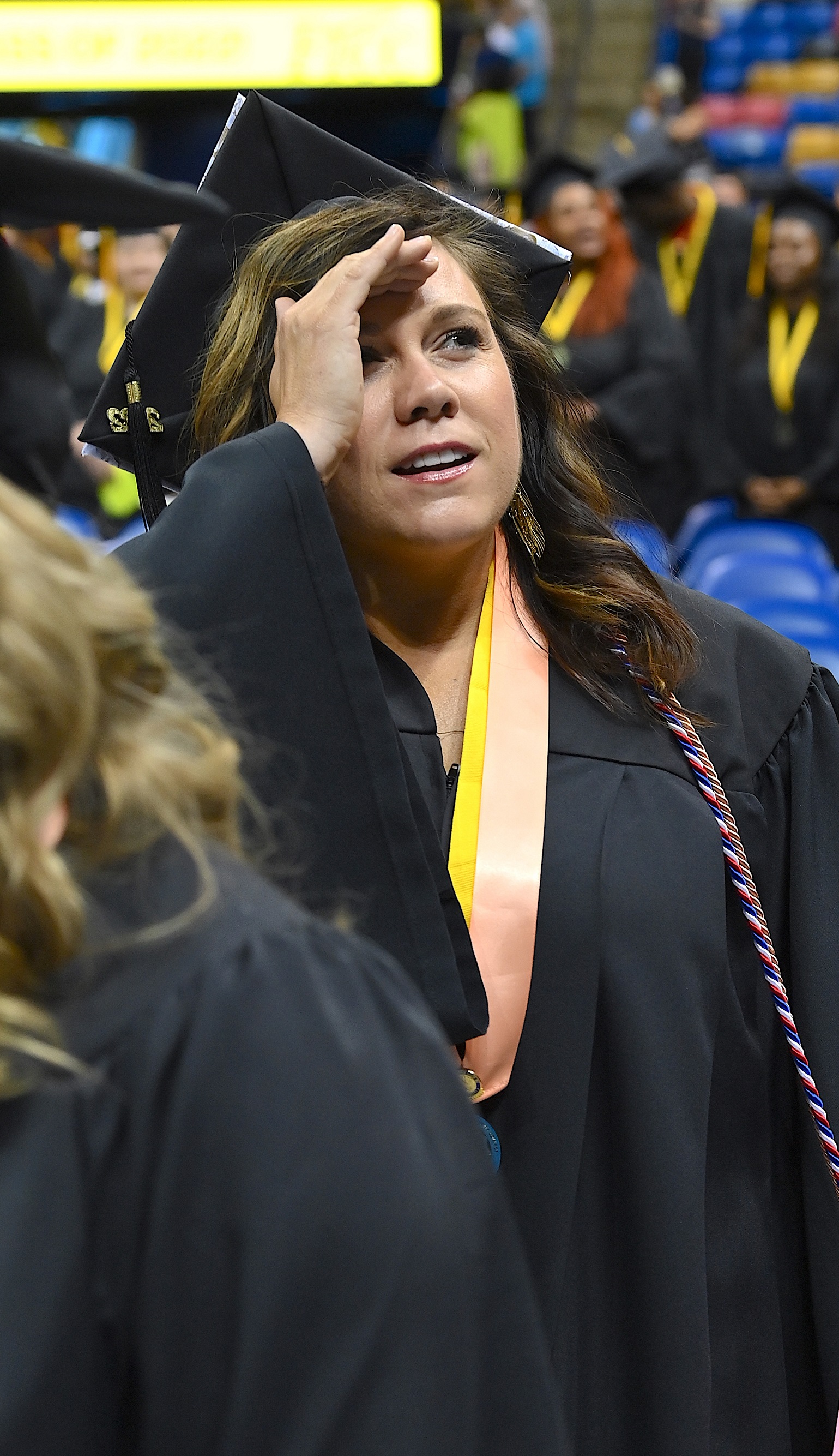 A graduate wearing a peach ribbon around her neck shields her eyes with her hand as she looks up into the crowd.