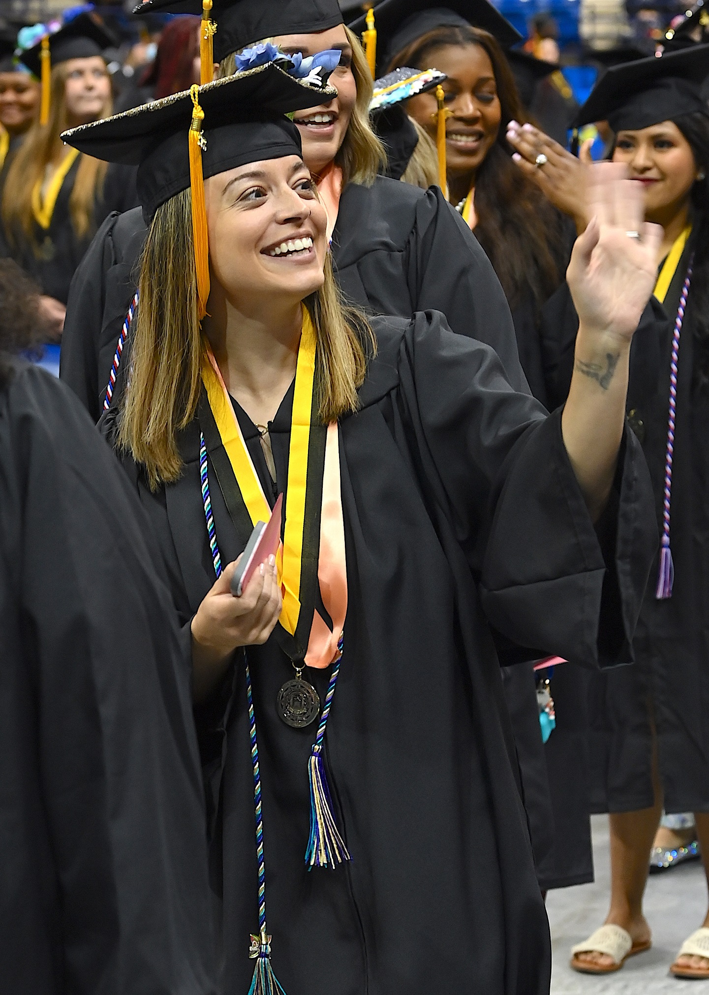 A graduate wearing a blue, white and teal cord and a medal on a black and gold ribbon waves to the crowd.