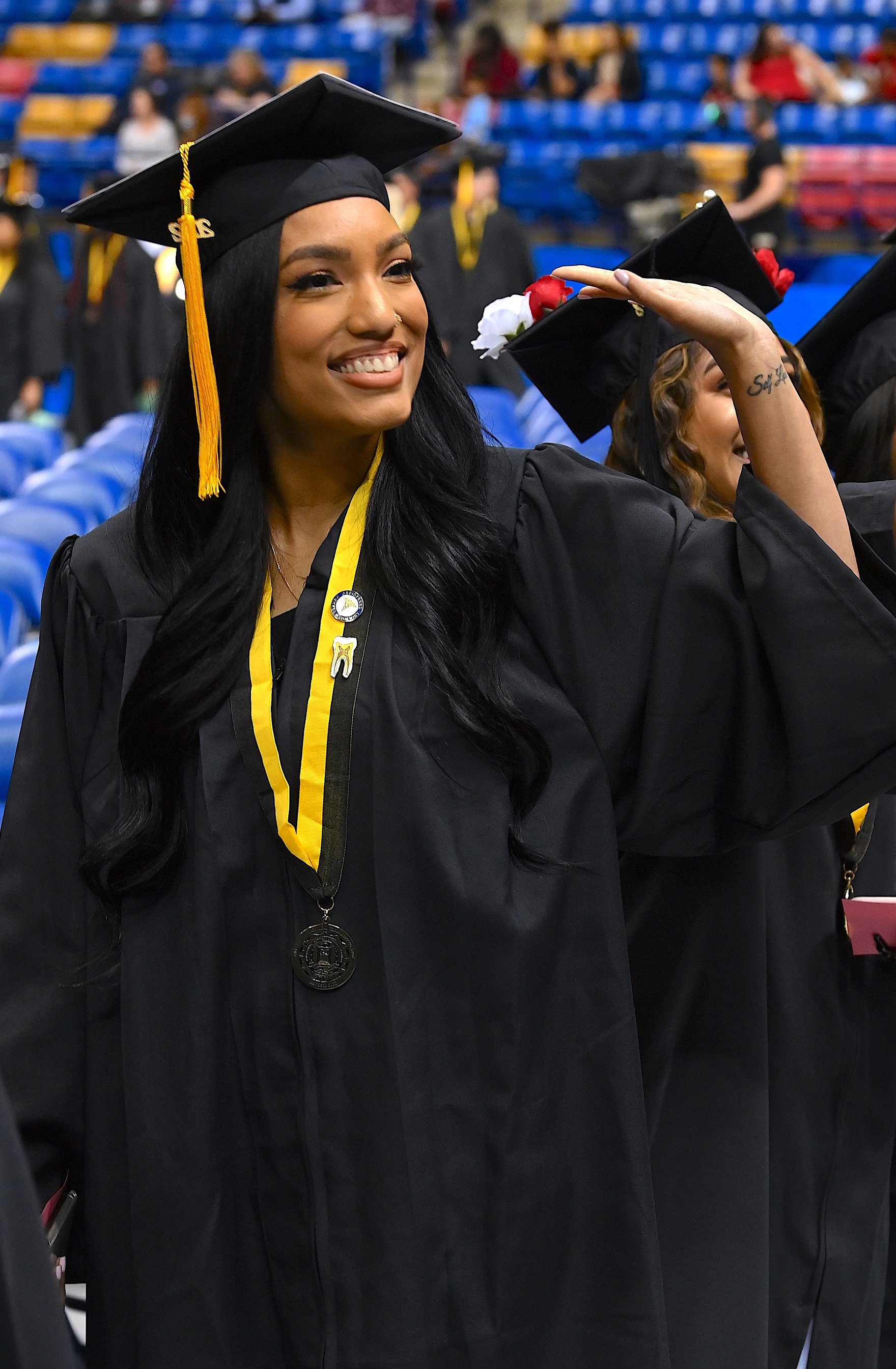 A graduate wearing a tooth enamel pin on her black-and-gold ribbon holding a medal around her neck waves to the crowd.