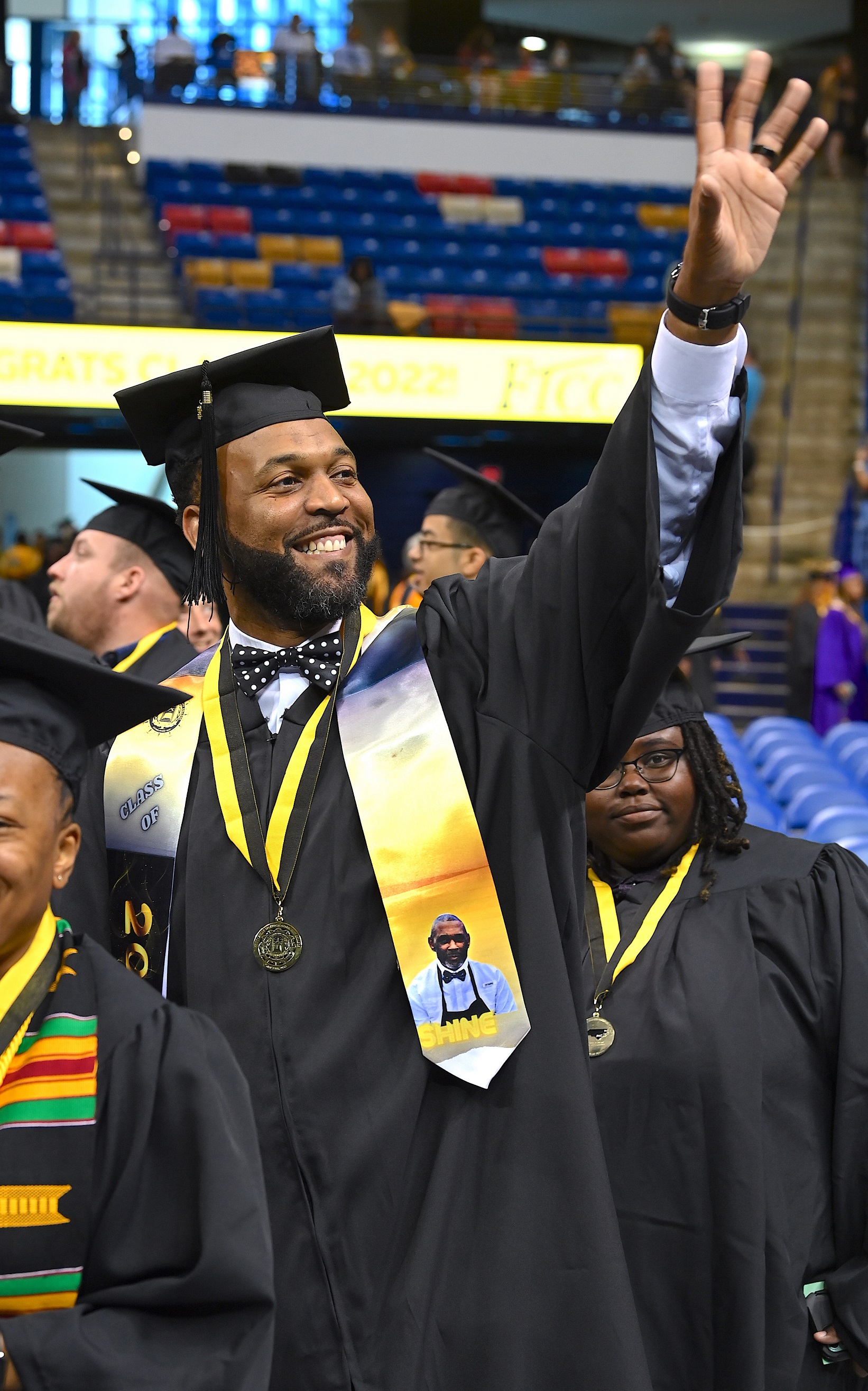 A graduate wearing a bow tie and a custom blue and gold stole raises his hand high to wave to the crowd.