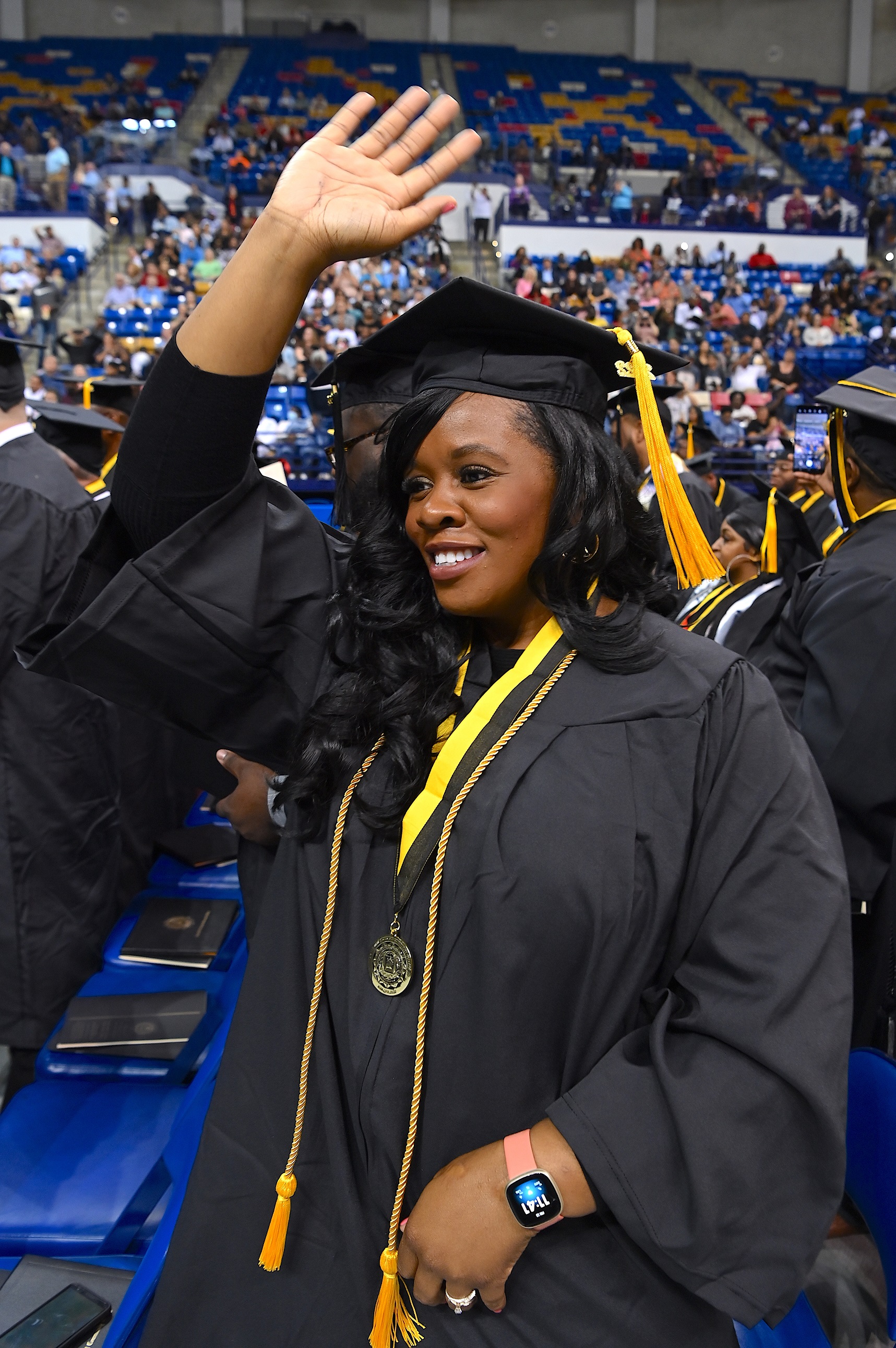 A graduate wearing a gold cord and a medal on a black-and-gold ribbon raises her hand to wave to the crowd.