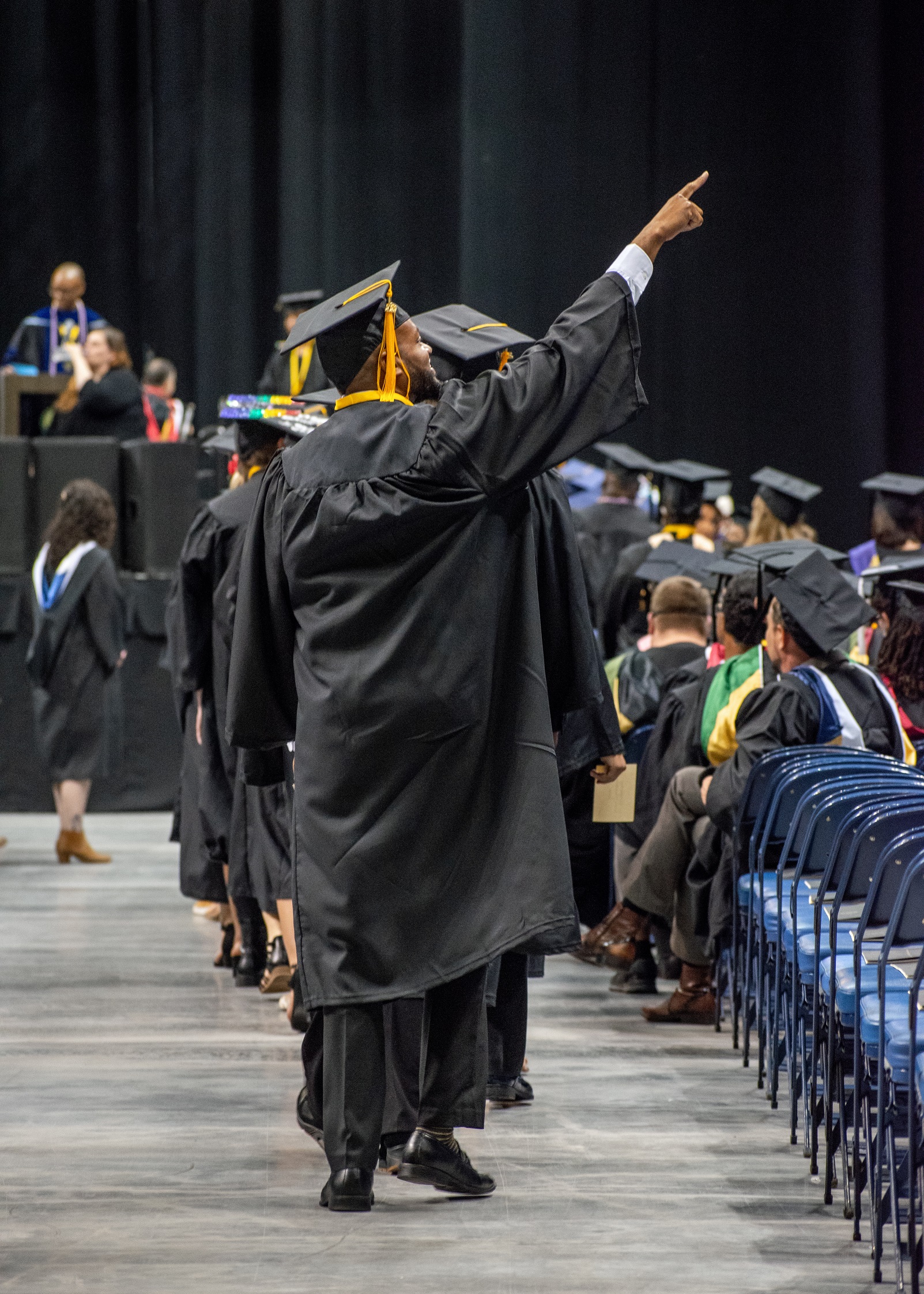 A graduate photographed from behind at the end of a line of graduates raises his arm high in the air to point up to the crowd.