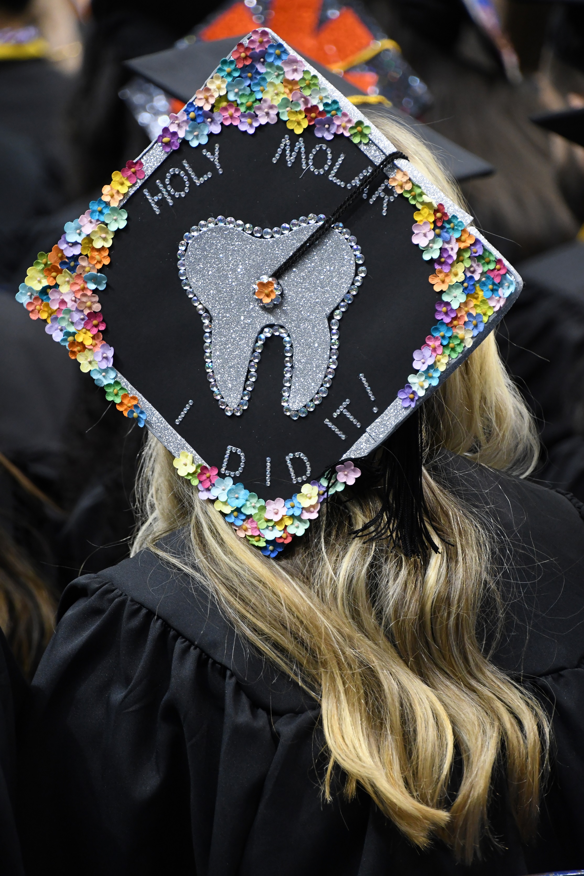 A graduation cap decorated with paper flowers and a silver paper tooth. The caps says "Holy Molar! I did it!"