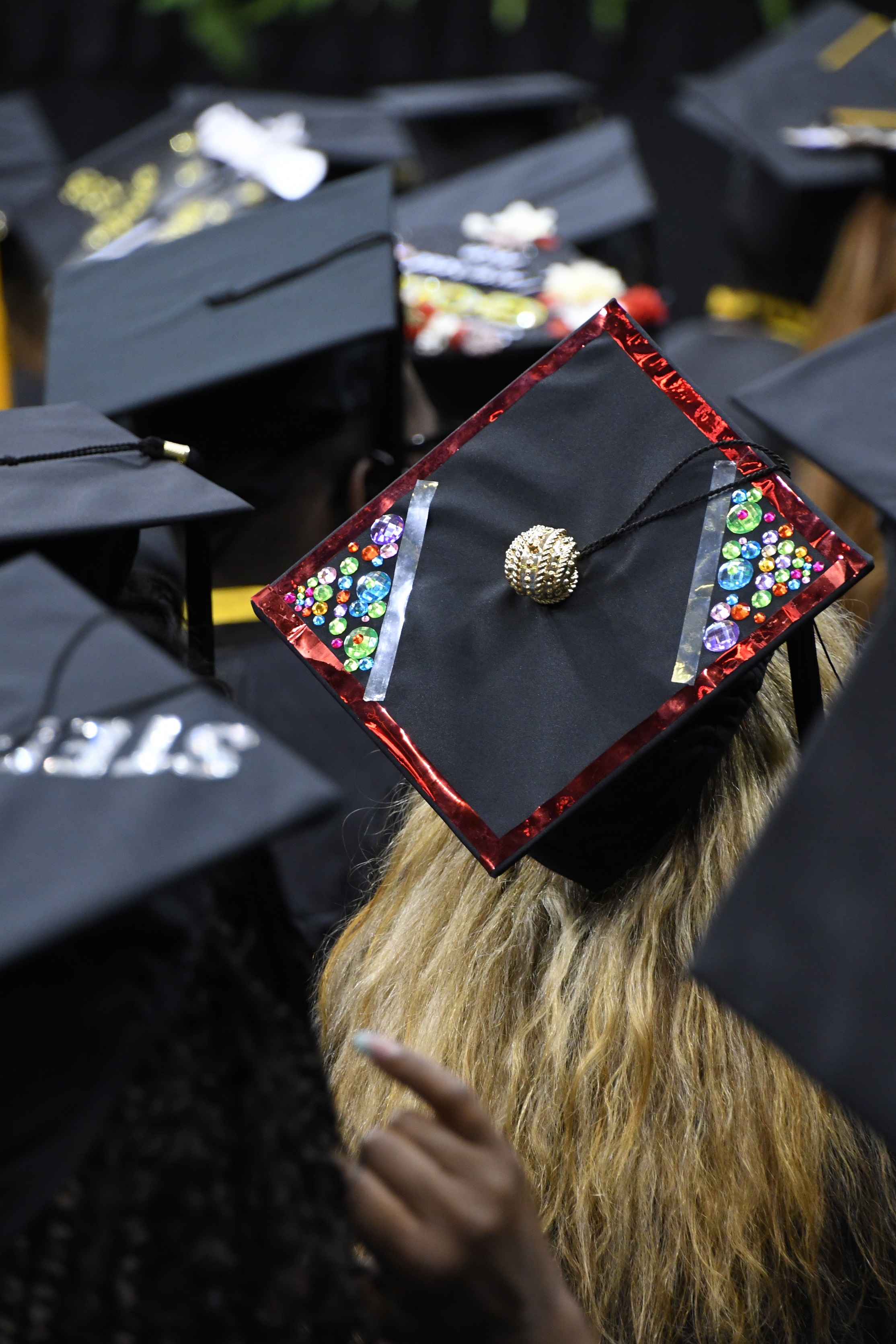 A graduation cap decorated with plastic gem stones, red trim and a gold topper