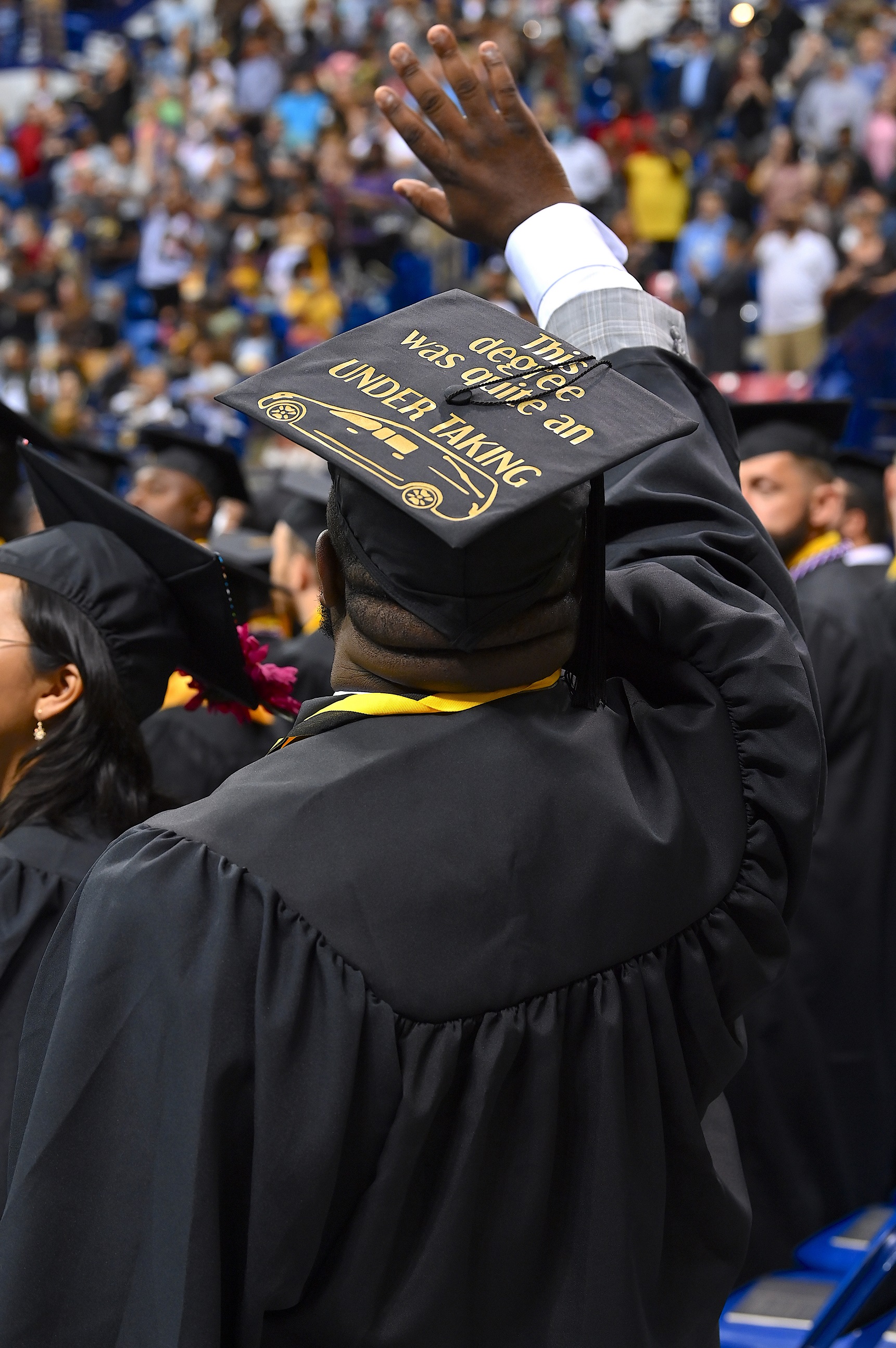 A graduate photographed from behind raises his hand in a wave. His cap says "This degree was quite an undertaking" and has a hearse drawn on it."