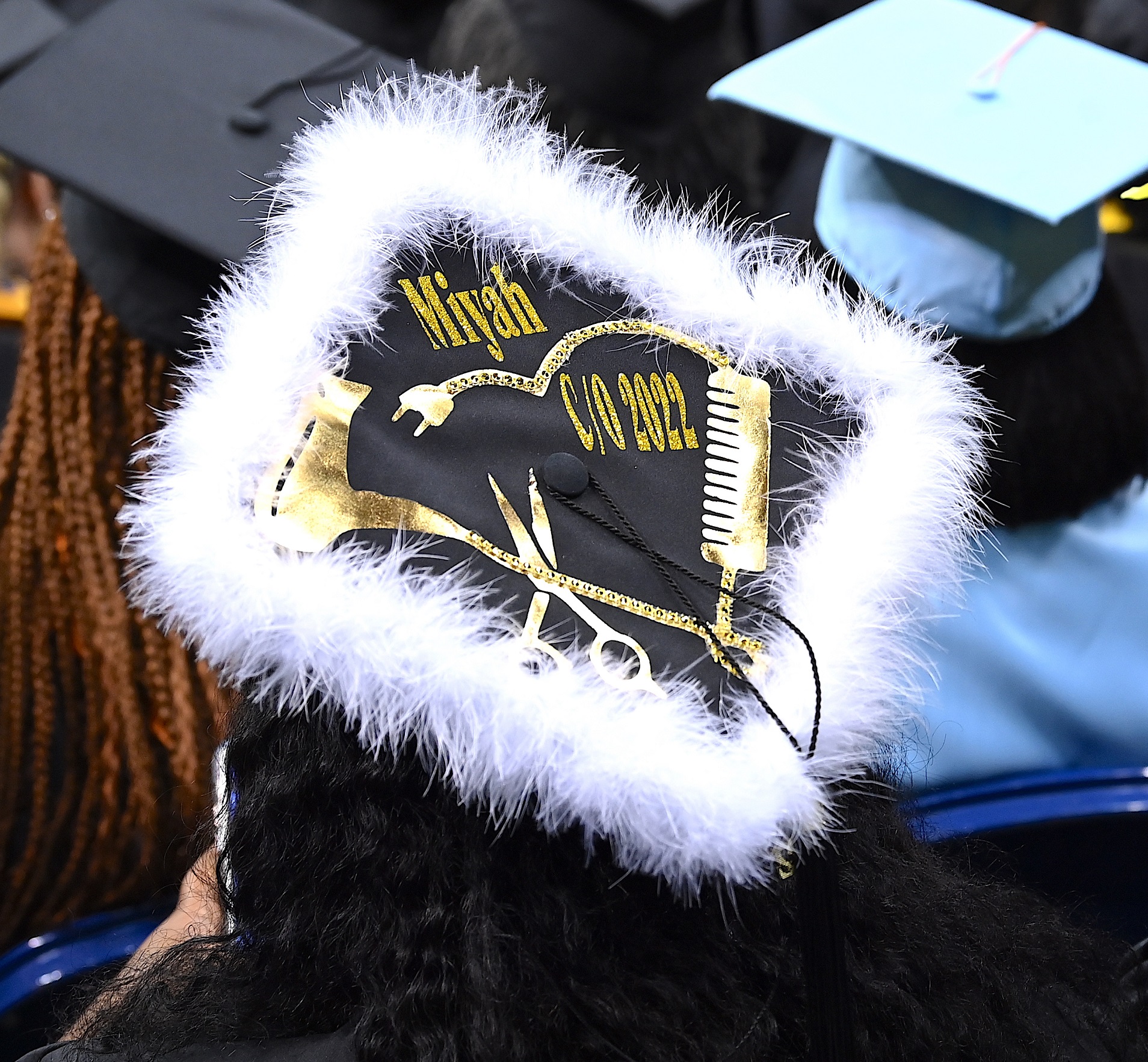 A graduation cap lined with white fur trim and a gold hair dryer, scissors and comb. The cap says "Miyah c/o 2022."