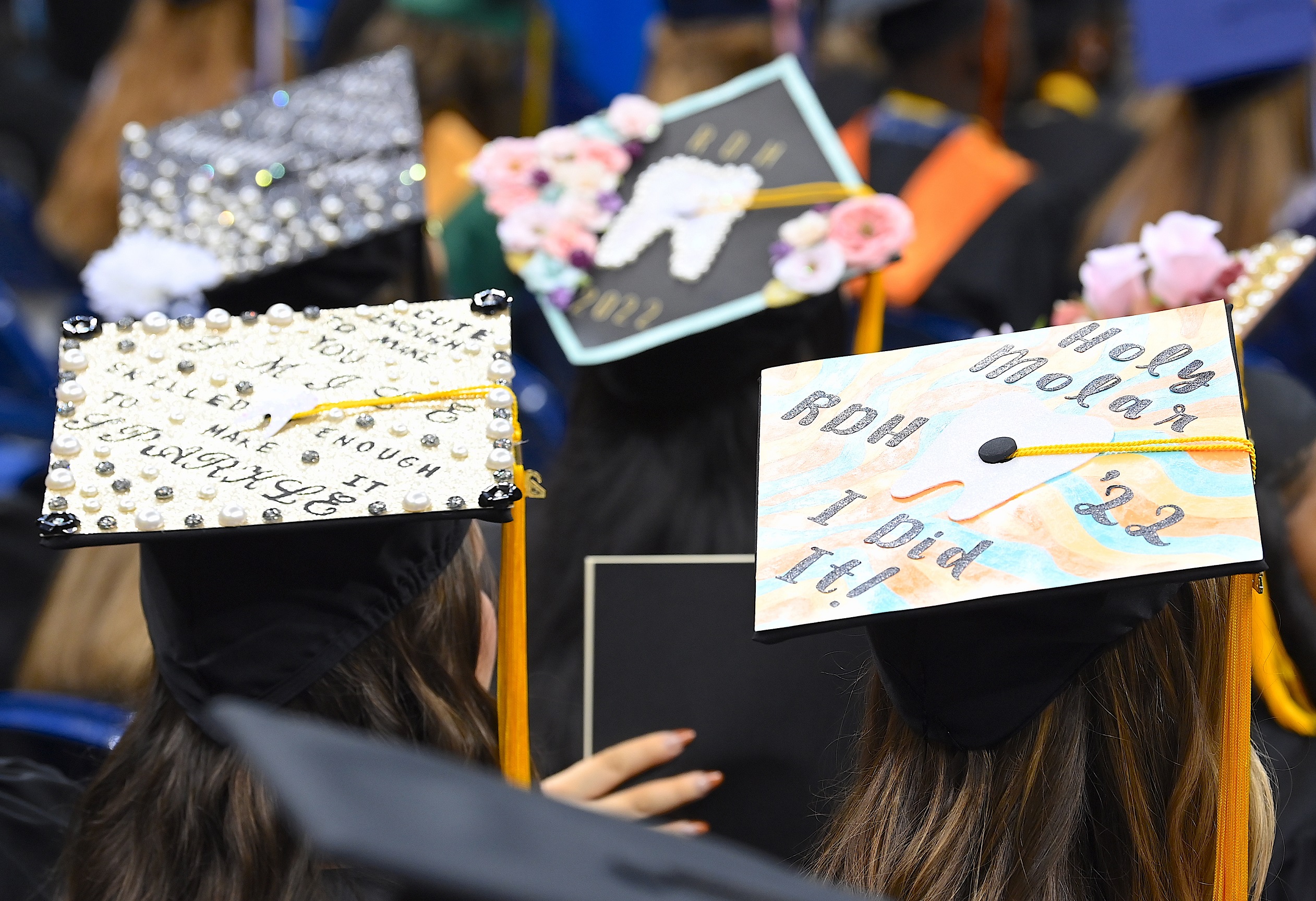Graduation caps in focus in the foreground of the photo. The one on the left says "Cute enough to make you smile. Skilled enough to make you sparkle." The one on the right says "Holy Molar. I did it! RDH '22"