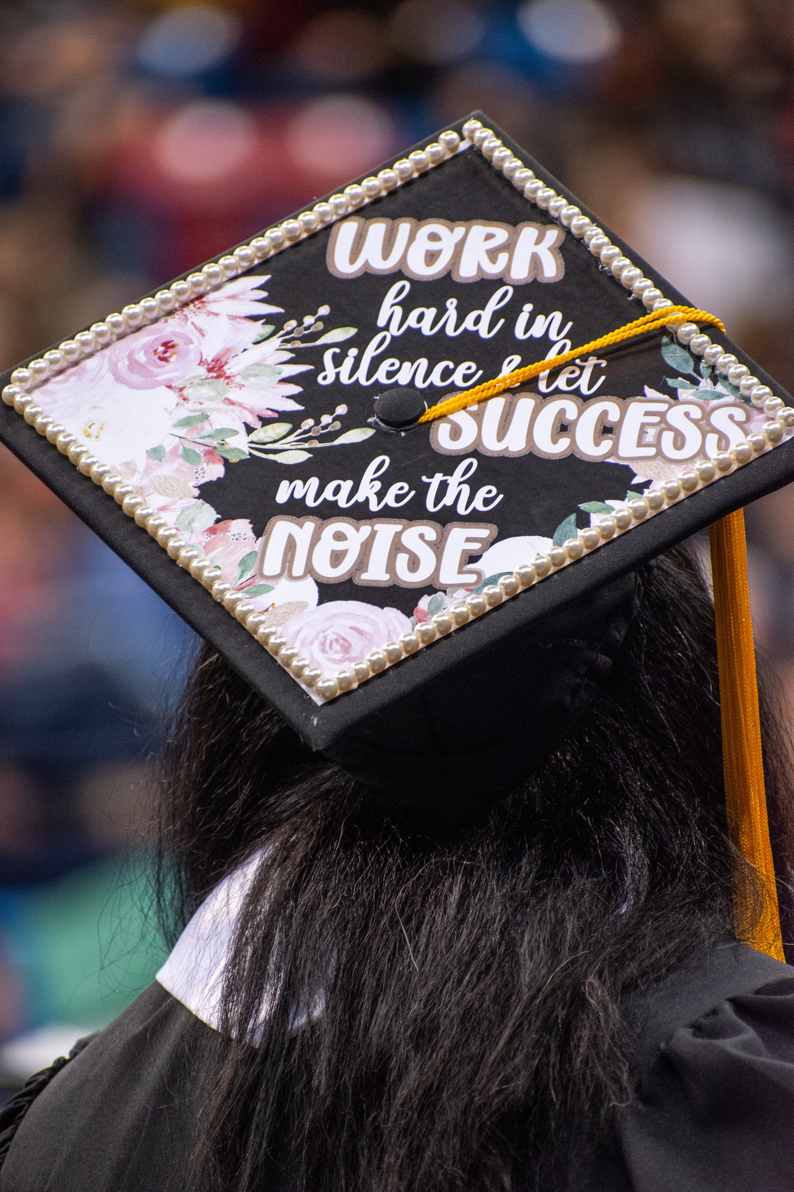 A graduation cap that says "Work hard in silence & let success make the noise." It is decorated with pearls and painted flowers.