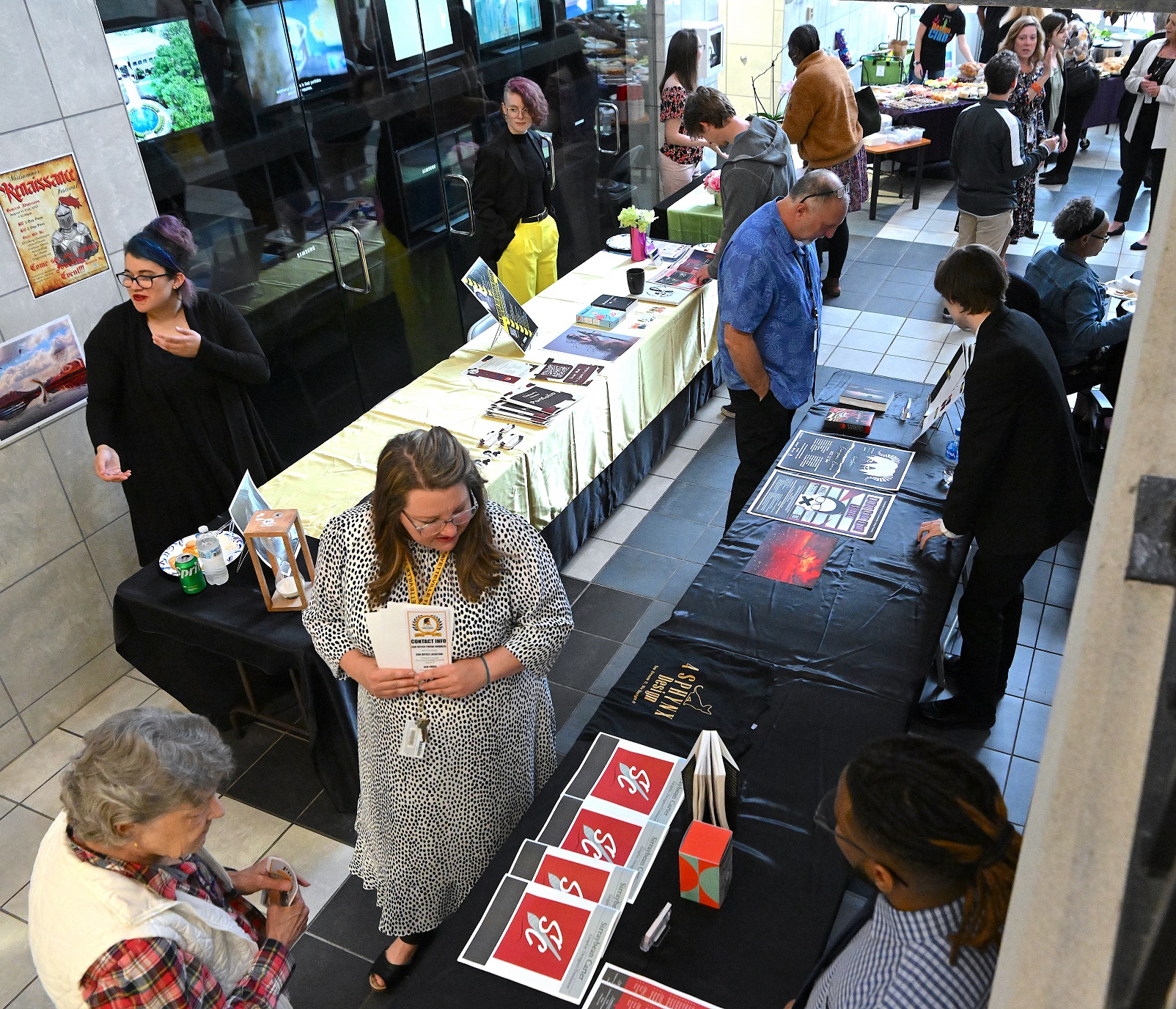 A wide photo of people moving through the row of tables where graduates display their portfolios and artwork.