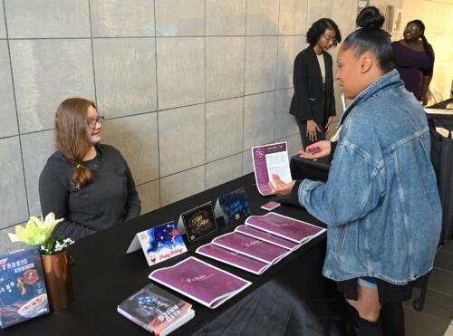 A woman stands at a table holding a business card while talking with a graduate who is seated behind the table.