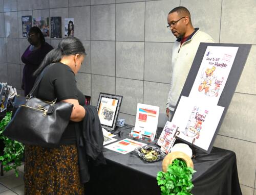A graduate stands behind a table and talks with a woman who is viewing his design work.