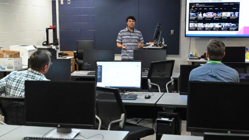 A graduate stands at the front of a computer lab to deliver a presentation of his digital media work.