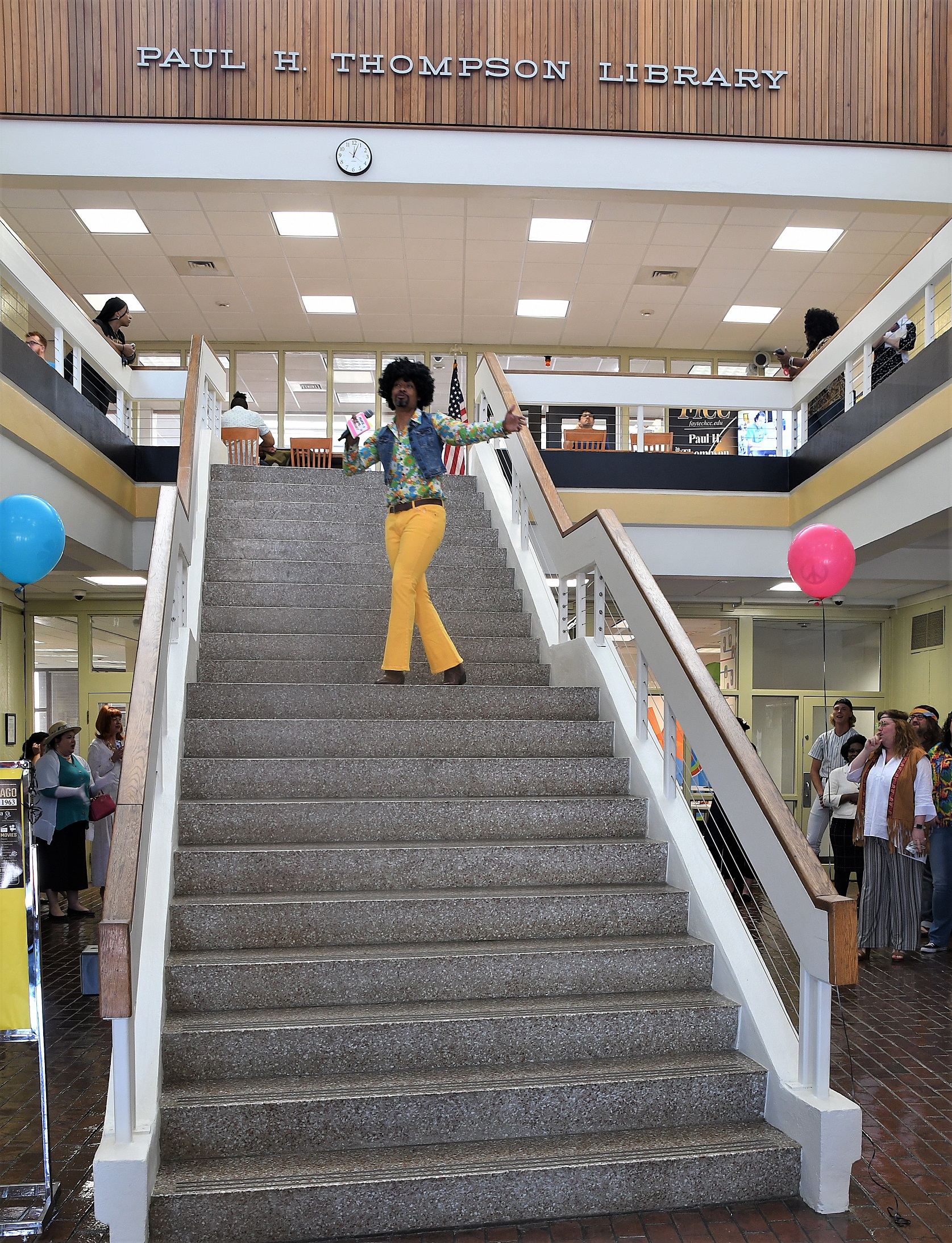 Costume contest host Sonny Kelly, dressed in bell-bottom jeans, a floral pattern shirt, denim vest and wig, stands on the steps in the library.