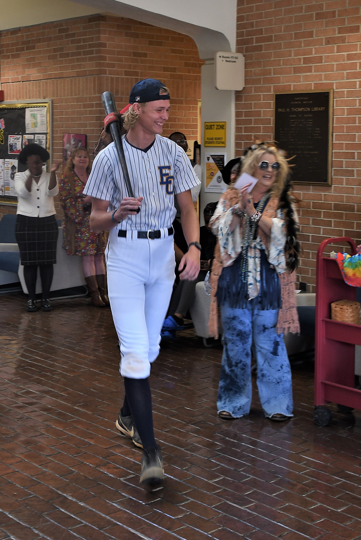 FTCC student Wyatt Barnaski, dressed as a 1960s-era baseball player and holding a baseball bat, walks to the center of the library lobby.