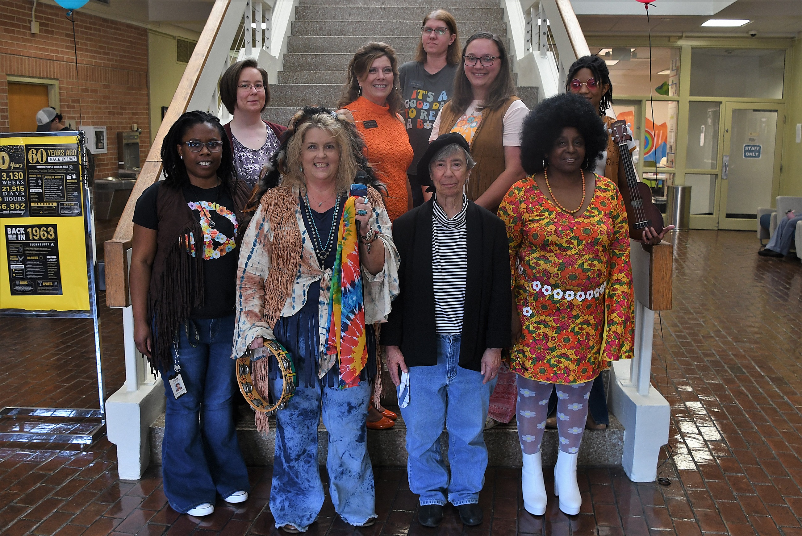 The library staff, dressed in 1960s-themed clothing, stand on the steps in the library lobby.