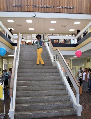 Costume contest host Sonny Kelly, dressed in bell-bottom jeans, a floral pattern shirt, denim vest and wig, stands on the steps in the library.