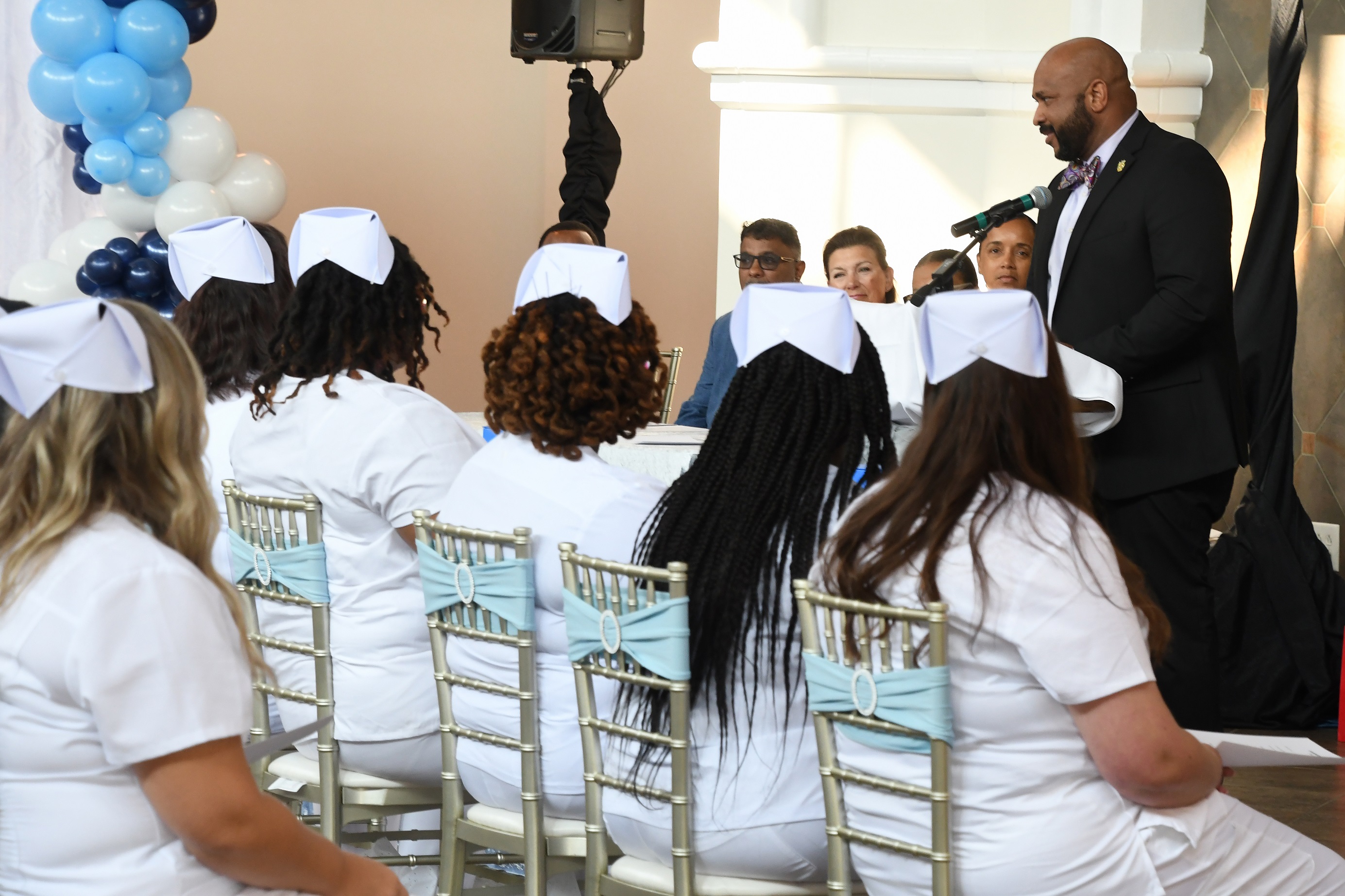 A guest speaker in a suit speaks at the microphone. The LPN graduates, seen from behind, are seated in front of him.